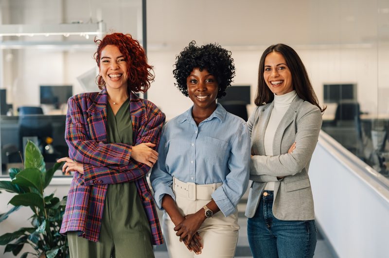 Drei lächelnde Frauen stehen nebeneinander in einem hellen Büro